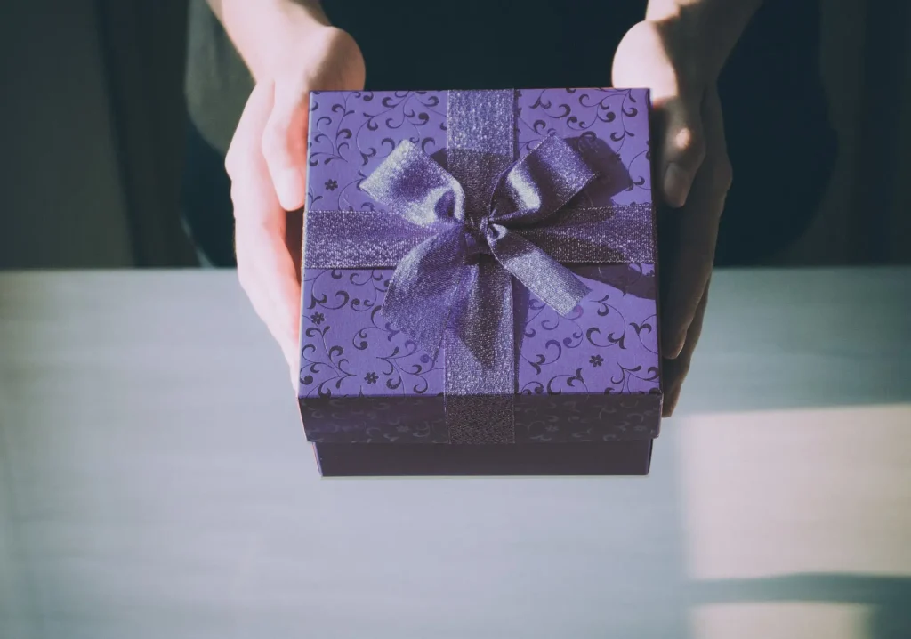 Mani che porgono una scatola regalo viola decorata con fiocco in tessuto brillante, illuminata da luce naturale su uno sfondo neutro