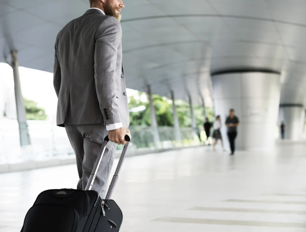 Persona in abito formale che cammina in un terminal moderno trascinando una valigia con rotelle.