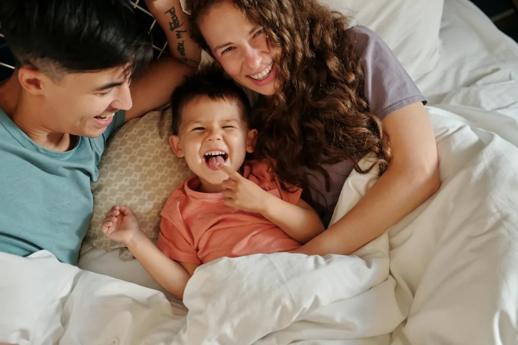 Persone riunite su un letto con coperte bianche, in un ambiente accogliente che richiama un soggiorno confortevole per famiglie.