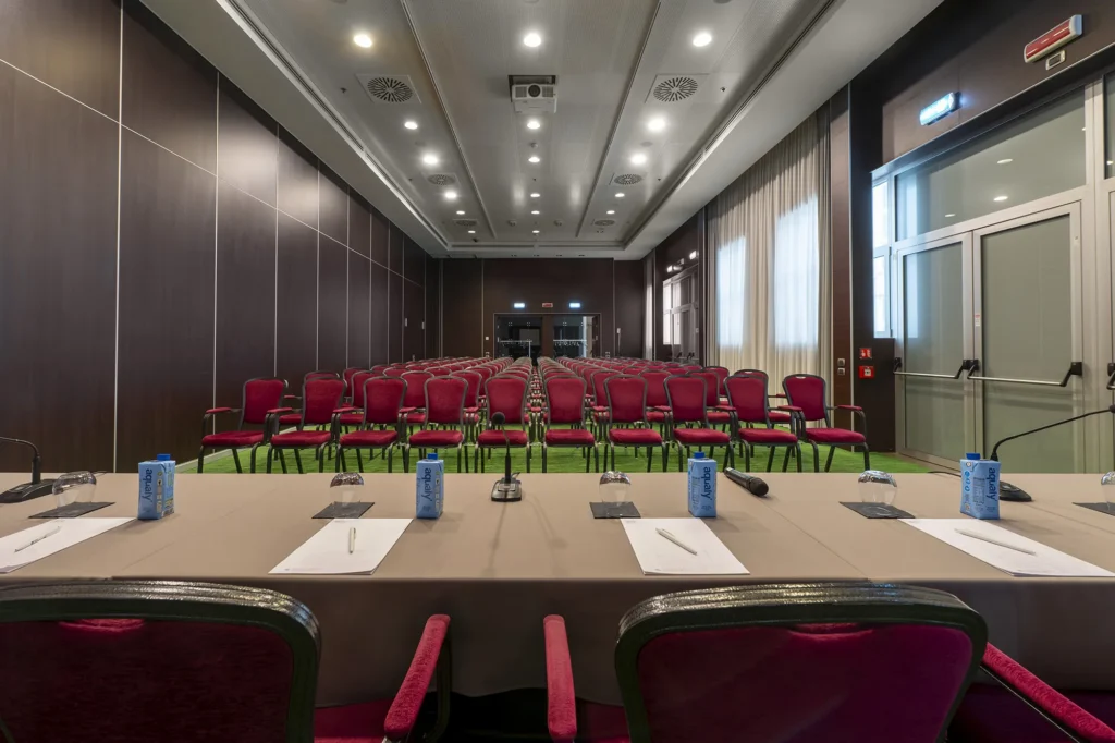 Sala meeting con disposizione a teatro, con file di sedie bordeaux orientate verso la zona relatori. In primo piano è presente un tavolo relatori con microfoni, blocchi appunti e bottiglie d’acqua. La sala ha moquette verde, pareti scure, ampie finestre laterali con tende e illuminazione a soffitto.