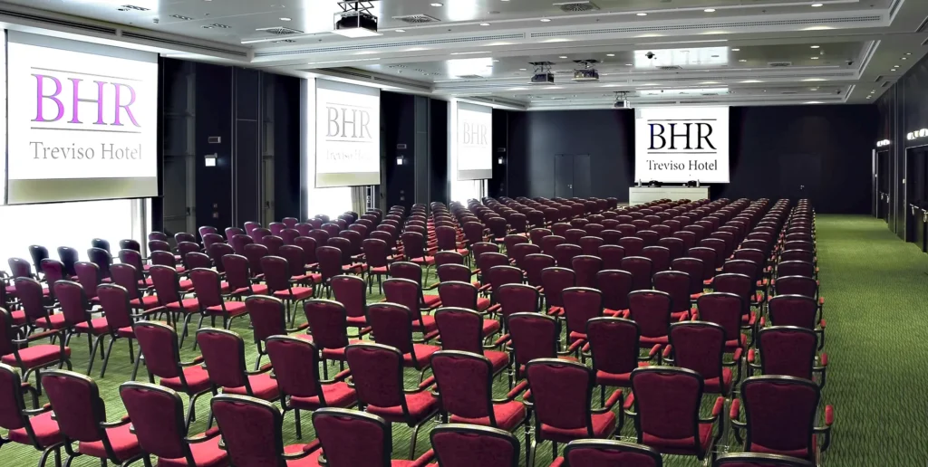 Sala conferenze ampia e luminosa con molte file di sedie rosse rivolte verso un palco con schermi che mostrano il logo ‘BHR Treviso Hotel’, pavimento in moquette verde e sala illuminata con faretti da soffitto.