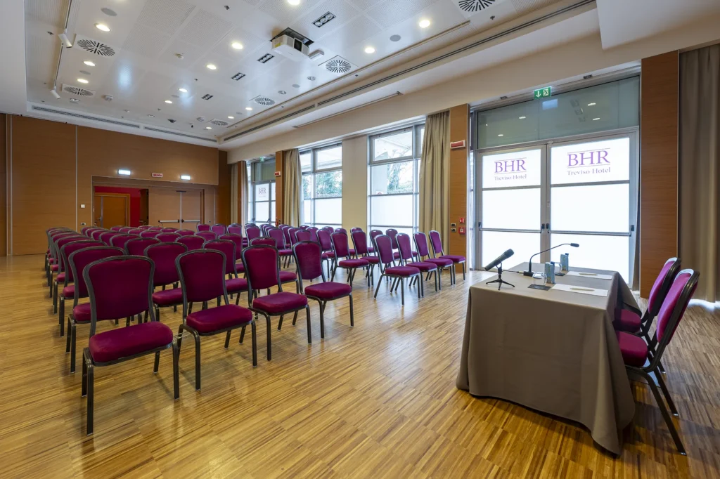 Sala meeting dell’hotel allestita a teatro, con file di sedie imbottite color bordeaux rivolte verso il tavolo relatori con microfoni. Ambiente ampio e moderno, con pavimento in legno, grandi finestre laterali, luci professionali a soffitto e pannelli “BHR Treviso Hotel” visibili sui vetri.