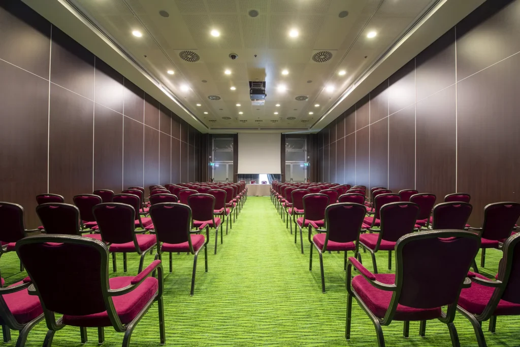 Sala meeting con disposizione a teatro, file di sedie viola imbottite rivolte verso il fronte. Tavolo relatori con microfoni e bottiglie d’acqua. Moquette verde, pareti scure e illuminazione a soffitto.