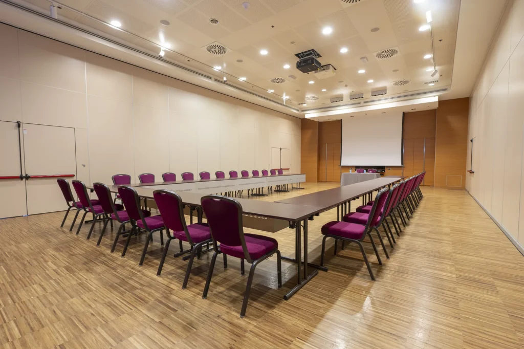 Sala meeting con disposizione a ferro di cavallo, tavoli rettangolari disposti su tre lati e sedie viola imbottite. Presente uno schermo per proiezioni. Pavimento in parquet, pareti chiare e illuminazione a soffitto creano un ambiente ordinato e professionale per riunioni e conferenze.