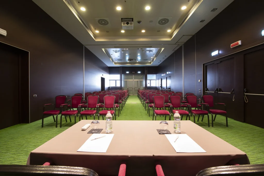 Vista frontale di una sala conferenze allestita in stile teatro con file di sedie imbottite color bordeaux su moquette verde. In primo piano un tavolo per relatori con bottiglie d’acqua e fogli; sullo sfondo porte d’ingresso, pareti scure, illuminazione a soffitto e proiettore centrale, ambiente professionale per eventi e meeting aziendali.