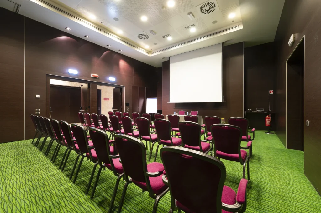 Sala conferenze moderna con sedie imbottite color bordeaux disposte in file su moquette verde, orientate verso uno schermo di proiezione bianco. Illuminazione a soffitto, pareti scure, tavolo relatori e lavagna a fogli mobili sul lato frontale.