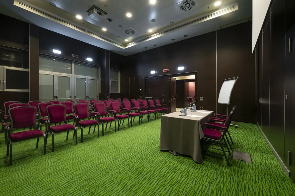 Sala conferenze ampia e luminosa con file di sedie rosse rivolte verso un palco con uno schermo e una lavagna a fogli mobili, pavimento in moquette verde e sala illuminata con faretti da soffitto.