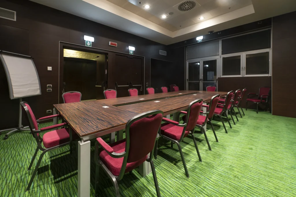 Sala riunioni con tavolo rettangolare in legno allestito per meeting. Attorno al tavolo sono disposte sedie imbottite rosse. Sul tavolo sono presenti bottiglie d’acqua e bicchieri. Sul fondo della sala si vede una lavagna a fogli mobili. Illuminazione con faretti da soffitto.