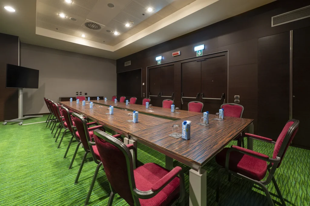Sala riunioni con tavolo rettangolare in legno allestito per meeting. Attorno al tavolo sono disposte sedie imbottite rosse. Sul tavolo sono presenti bottiglie d’acqua e bicchieri. Sul fondo della sala si vede un monitor a schermo piatto. Illuminazione con faretti da soffitto.