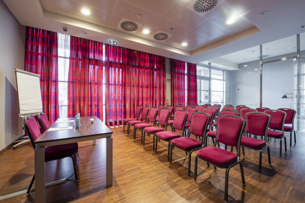 Sala meeting dell’hotel allestita a teatro con file di sedie rosse, tavolo relatori con lavagna a fogli mobili, pavimento in legno e grandi tende rosse filtranti la luce naturale.