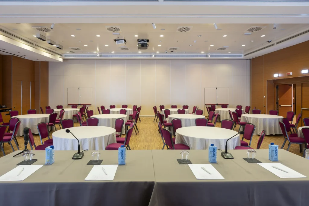 Ampia sala conferenze con allestimento a cabaret, tavoli rotondi con sedie bordeaux, tavolo relatori con microfoni e blocchi appunti, pavimento in legno e illuminazione a soffitto.