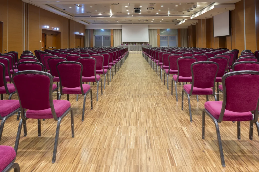 Sala meeting dell’hotel allestita a platea, con sedie imbottite color bordeaux disposte in file ordinate e corridoio centrale, rivolte verso il palco con tavolo relatori, podio e schermo di proiezione. Ambiente ampio e professionale, con pavimento in legno, pareti in legno e pannelli chiari, illuminazione tecnica a soffitto e porte laterali con vetri brandizzati “BHR Treviso Hotel”.